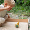 A child in light attire squats by a sand-filled wooden tray, intrigued by a small spinning top. Nearby, the Grapat Flowing Yellow Bird Rocking Toy flits playfully amidst the blurred greenery.