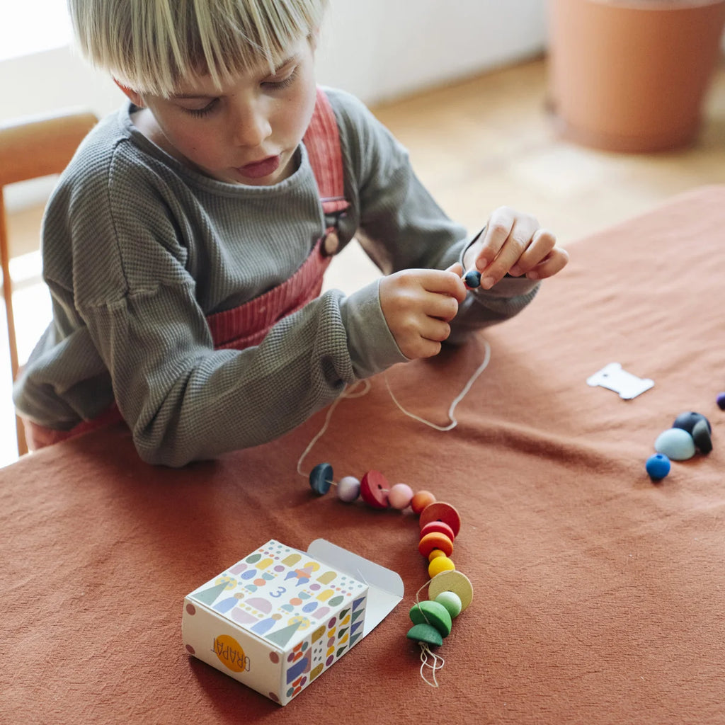 A child threads colorful wooden beads onto a string at a table covered with a rust-colored cloth, while an open box with patterns—part of the Grapat Christmas Advent Calendar 2026 (Final Sale, ships in approximately two weeks)—sits nearby, sparking creative family holiday activities.