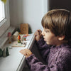 A young child in a purple sweater looks intently at a miniature toy scene arranged on a windowsill, featuring tiny trees, a gnome, a lamp, and other small objects—all part of the Grapat Christmas Advent Calendar 2026 – Final Sale (ships in approximately two weeks), perfect for family holiday activities.