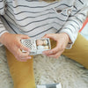A child sits cross-legged on a fuzzy rug, holding an open box from the Grapat Christmas Advent Calendar 2026 - Final Sale (ships in approximately two weeks), revealing four small, speckled egg-shaped objects inside. Wearing a striped shirt and mustard yellow pants, the child is ready to enjoy special family holiday activities.