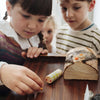 A young child places a small wooden piece near a toy log and chest, while two other children watch attentively in the background—capturing a heartwarming moment of family holiday activities inspired by the Grapat Christmas Advent Calendar 2026.