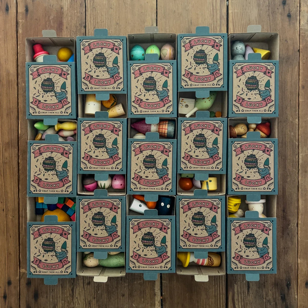Sixteen small boxes from the Grapat Lucky Lucky Surprise Set 5th Edition are arranged in a grid on a wooden floor. Each mystery box is partly open, revealing an assortment of colorful collectible toys and objects inside each compartment.