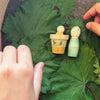 Two small wooden dolls from the Grapat Lucky Lucky Surprise Set 5th Edition rest in a yellow cup atop large green leaves. A child’s hands reach eagerly toward the dolls, ready to discover the playful treasures hidden inside this charming collectible toy set.