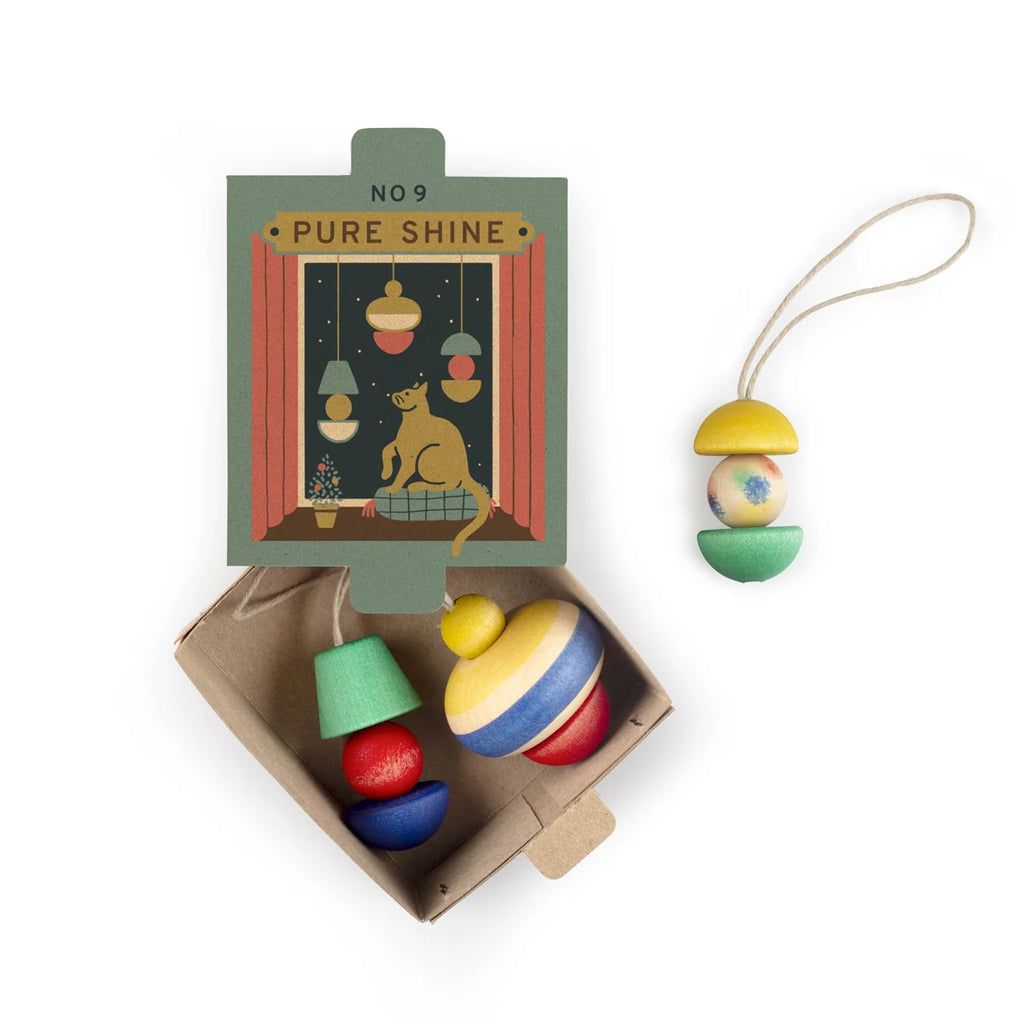 A set of colorful, hand-painted wooden ornaments from Grapat Ornament Set - No 9 - Pure Shine sits next to a box featuring a cat and lights; one ornament hangs from a string, others rest in an open cardboard tray.