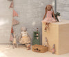 A Maileg Bunny, Size 3 - Classic - Ballet Suit and Skirt - Cream, is posed by a wooden block, with teacups, suitcase, potted plant, green shelf, and bunting flags in the softly lit background for a whimsical ballet-themed scene.