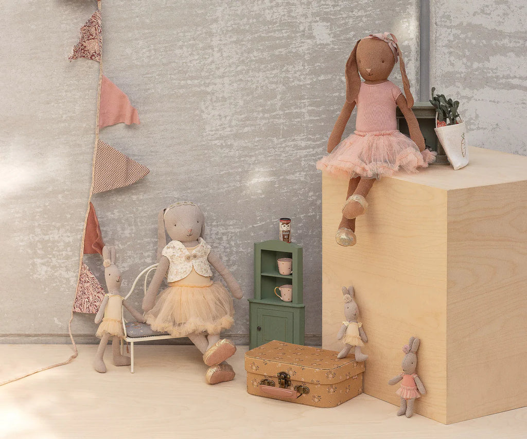 A Maileg Bunny, Size 3 - Classic - Ballet Suit and Skirt - Cream, is posed by a wooden block, with teacups, suitcase, potted plant, green shelf, and bunting flags in the softly lit background for a whimsical ballet-themed scene.