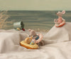 Two Maileg toy mice sit on fabric dunes at the beach. One relaxes on the Maileg Rubber Boat, Small Mouse - Yellow Stripe, while the other sits higher up on a floral ring. A toy bucket and sand shovel are nearby.