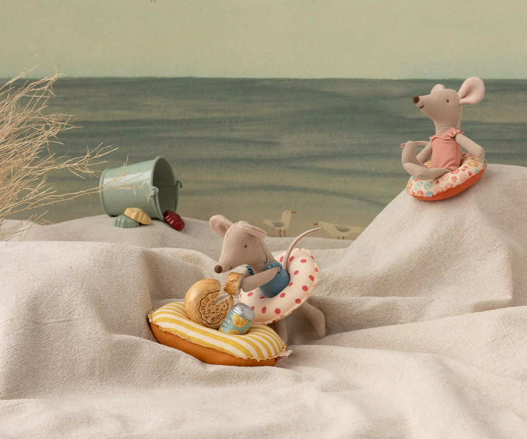 Two Maileg toy mice sit on fabric dunes at the beach. One relaxes on the Maileg Rubber Boat, Small Mouse - Yellow Stripe, while the other sits higher up on a floral ring. A toy bucket and sand shovel are nearby.