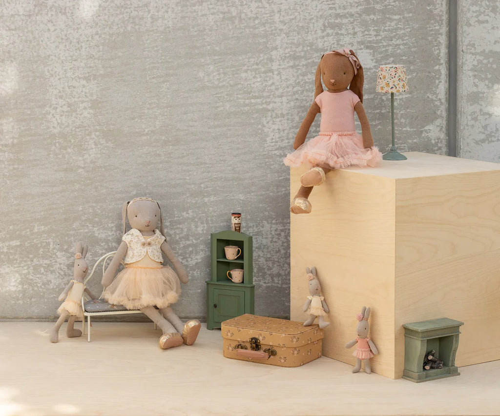 A Maileg Bunny, Size 3, in chocolate brown with a ballet suit and rose skirt sits among miniature furniture—green cabinet, small suitcases, lamp, and bench—against a neutral background.