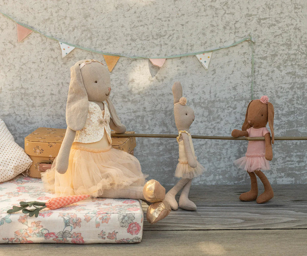 A Maileg Bunny, Size 1, in chocolate brown wears a rose ballet suit and skirt, posed like a dancer on a floral mattress with suitcases behind and pastel bunting on the textured wall.