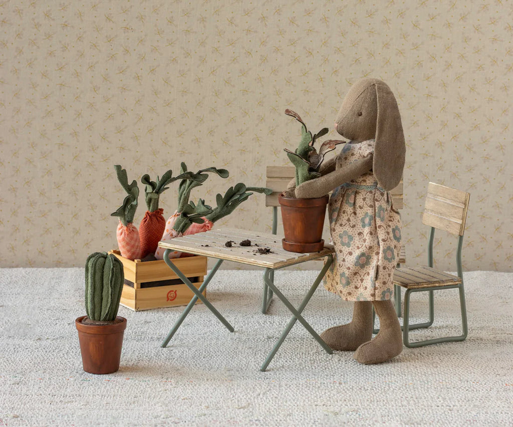 The Maileg Bunny, Size 2 - Brown - Dress stands by a small table, repotting a fabric plant. Soft natural fabrics form nearby plants and veggies, with tiny furniture and a crate in the background.