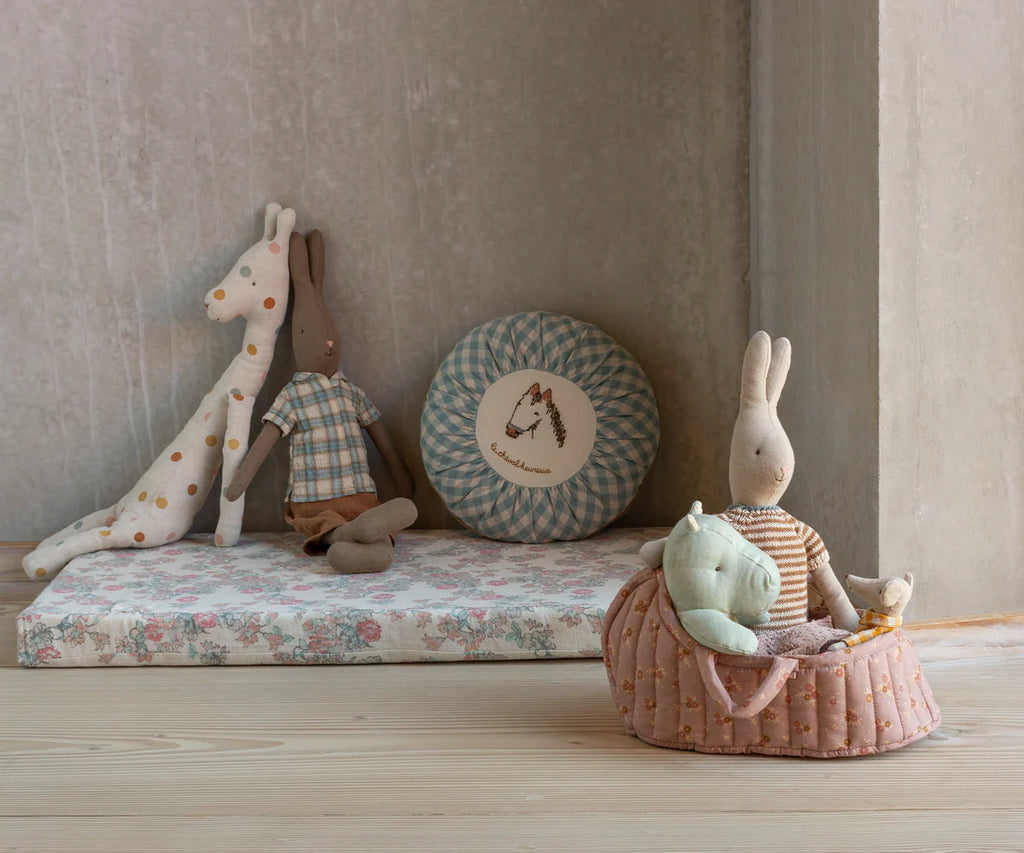 A cozy children's play area features plush toys—a giraffe, two striped rabbits, a bunny bed, a mountain cushion—and the Maileg Carry Cot, Large - Rose as a pink basket holding stuffed animals, all atop a wooden floor.