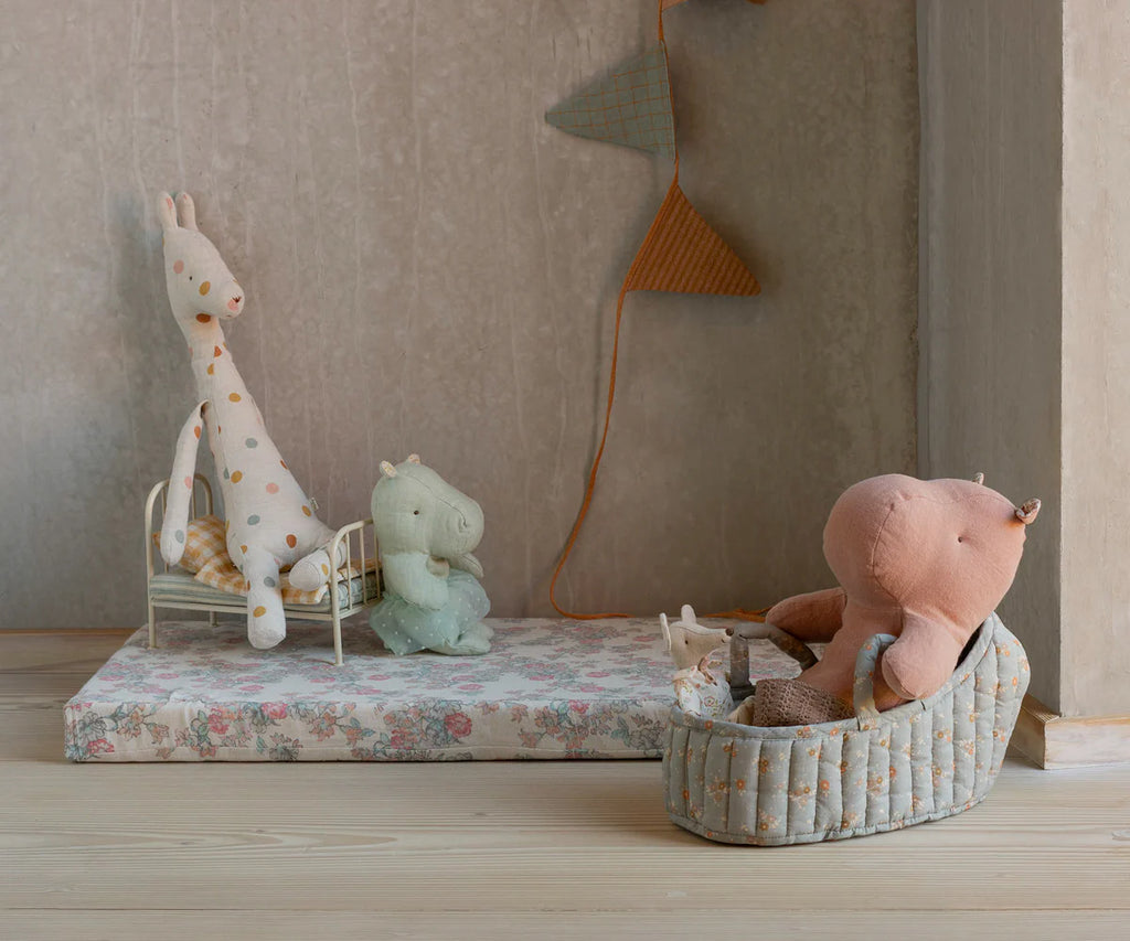 A soft toy giraffe and two hippos rest on a bed and in the Maileg Carry Cot, Large - Blue with toy bedding, atop a floral mattress in a cozy, neutral-toned room decorated with fabric pennant garlands.