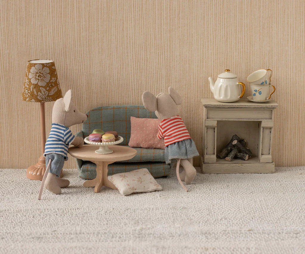 Two toy mice in striped shirts stand by the Maileg Oval Coffee Table, Mouse - Nature, with pastries. One mouse leans on the sofa, the other stands nearby. A lamp, fireplace, tea set, and cushion finish this cozy mouse-size SuitCasa scene.