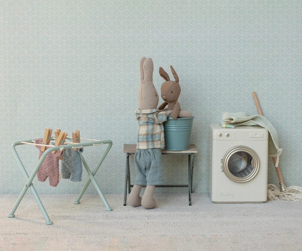 Two stuffed rabbits are in a pastel laundry room; one places the smaller rabbit, dressed in a Maileg Extra Clothing: Knitted Suit, Micro - Blue for Micro Bunny, into a blue bucket. Nearby are a toy washing machine and miniature clothes on a drying rack.