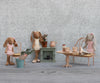 Three fabric bunny dolls in pink outfits sit in a cozy miniature home with a table, chairs, potted plants, and the Maileg Fireplace, Miniature - Dusty Green—all set against a gray textured wall background.