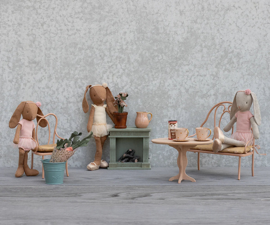 Three fabric bunny dolls in pastel dresses gather around the Maileg Vintage Bench, Mini - Dark Powder, along with miniature furniture pieces like a tea table, fireplace, and potted plant against a gray textured wall.