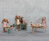Three fabric bunny dolls in pink clothes have a mini tea party with Maileg’s Vintage Chair, Mini - Dark Powder, surrounded by tiny rabbit furniture, teacups, a plant, and a decorative fireplace against a textured gray wall.