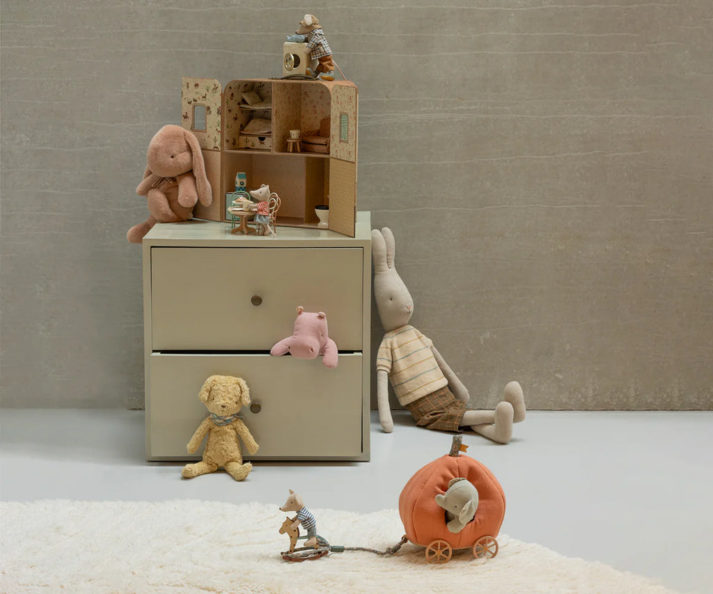 A small dresser with two drawers holds a dollhouse and stuffed animals, including the Maileg Plush Bunny, Medium - Vintage Rose, plus a bear and mouse. More plush toys and a pumpkin-shaped carriage sit on the floor by a white rug against a gray wall.