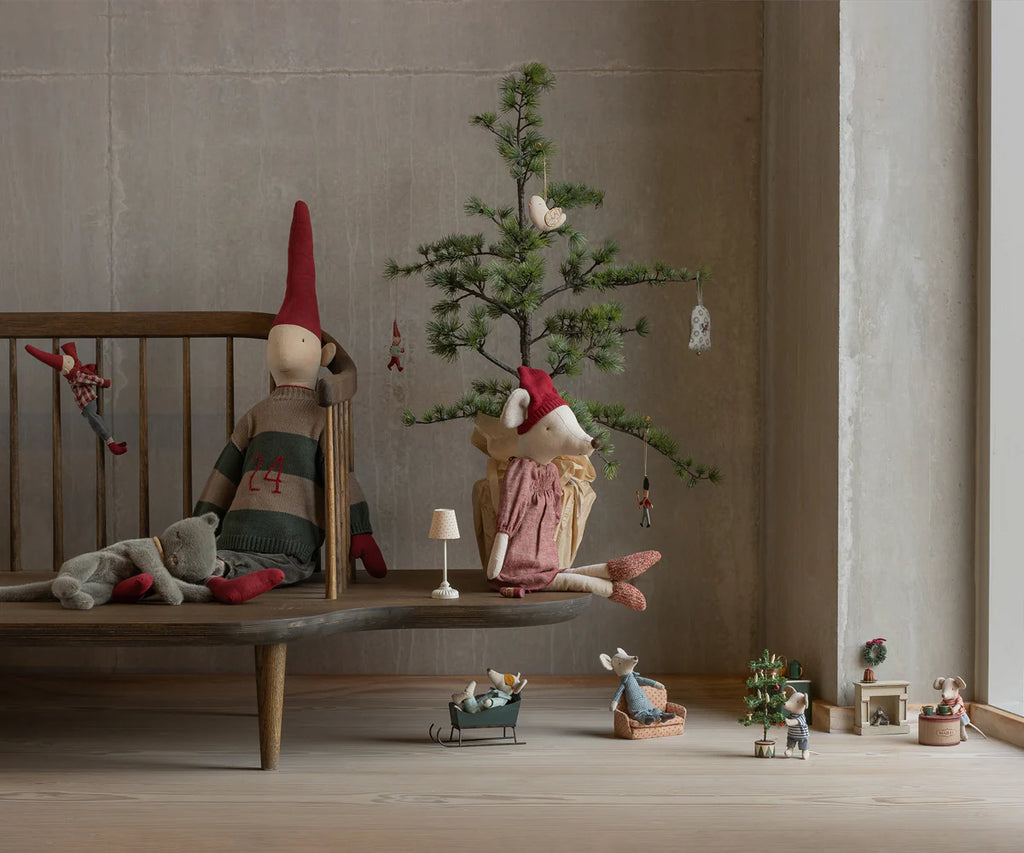 A Maileg Christmas Mouse, Maxi - Girl sits on a wooden bench by a small decorated tree, surrounded by miniature furniture and festive holiday decorations. Ships in one week.