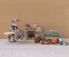 Two toy mice in a cozy miniature living room: one mouse irons doll clothes while the other packs a suitcase. The scene features Maileg Extra Clothing—Jacket, Pants, Hat, and Bow Tie for Grandpa Mouse—plus sofa, fireplace, food, and more.