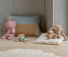 The Maileg Plush Bunny, Medium - Cream Peach sits on a light wood floor alongside a pink hippo plush and a small doll on a polka-dot cushion, with a pillow and cream rug against a concrete wall.