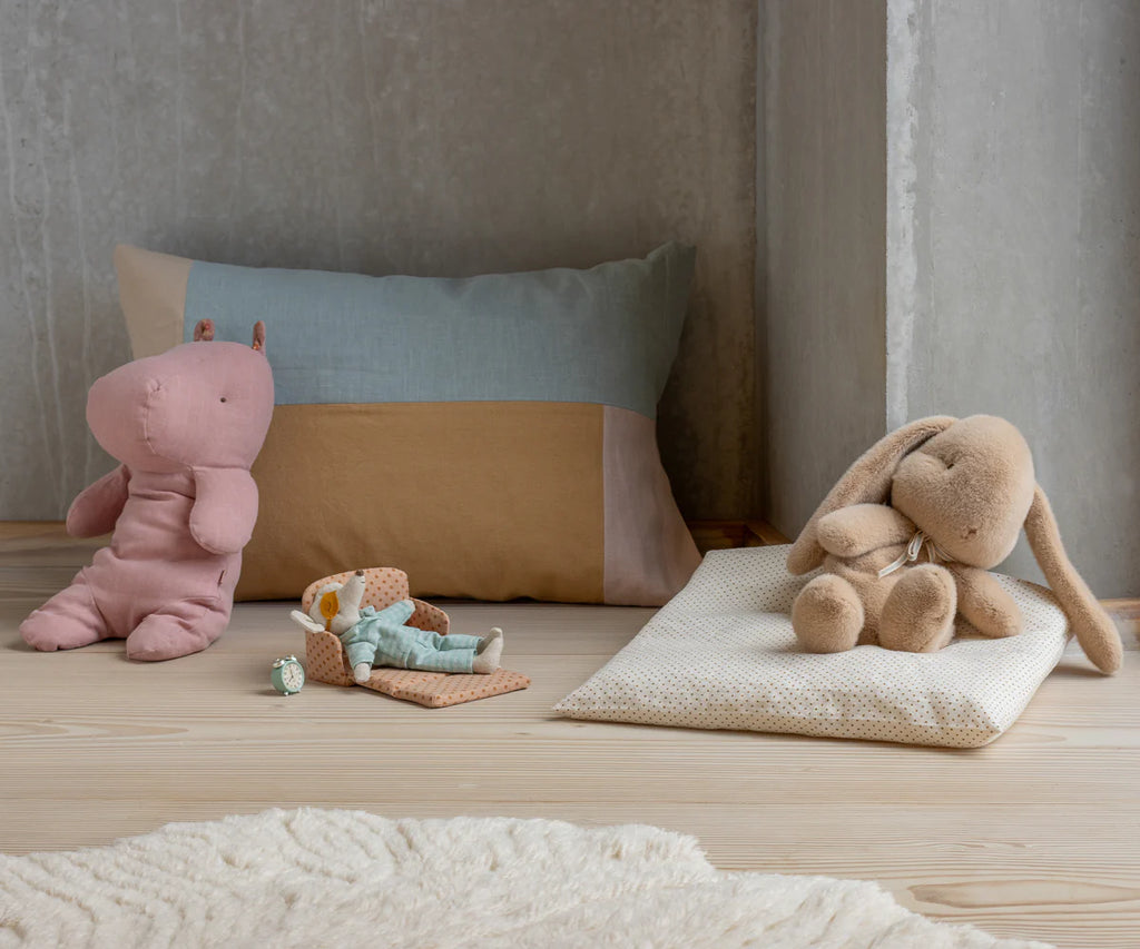 The Maileg Plush Bunny, Medium - Cream Peach sits on a light wood floor alongside a pink hippo plush and a small doll on a polka-dot cushion, with a pillow and cream rug against a concrete wall.