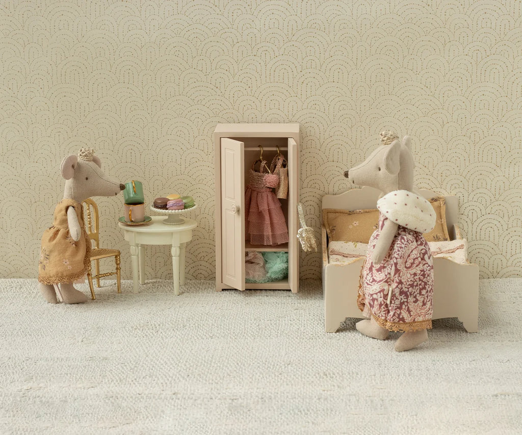 Three toy mice, dressed in clothes, are in a miniature room with a bed, a Maileg Mini Side Table topped with pastries, a chair, and an open wardrobe displaying hanging outfits. The scene features a neutral, vintage style.