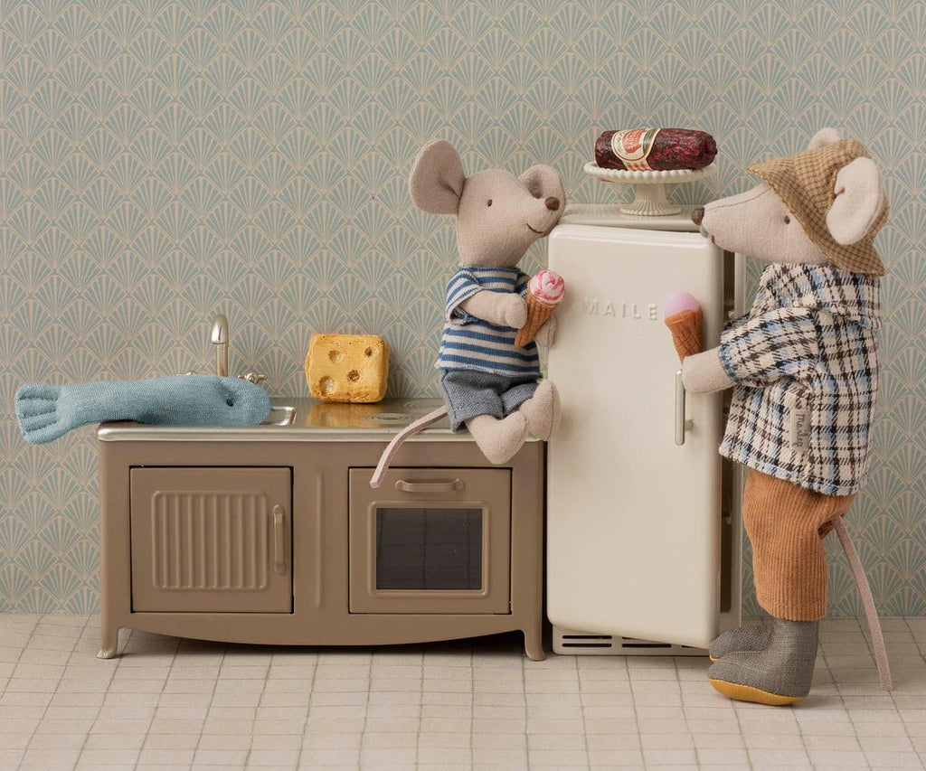 Two toy mice in a miniature kitchen, one dressed in Maileg Extra Clothing: Jacket, Pants, Hat and Bow Tie for Grandpa Mouse, enjoy ice cream with fridge, cheese, fish, patterned wallpaper, and charming doll clothing details.