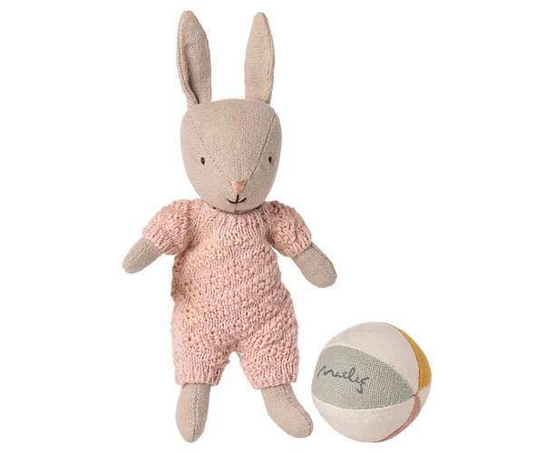 The Maileg Rabbit With Accessories, Micro - Sand - Rose stands beside a pastel fabric ball labeled "Maileg," both set against a plain white background. The rabbit wears a pink knitted outfit, making it a charming storytelling toy.