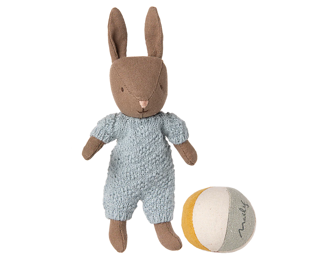 The Maileg Micro Bunny With Ball - Brown - Blue (With Magnetic Hands) wears a light blue knitted outfit and stands next to a small soft ball with white, yellow, and gray panels on a plain background.