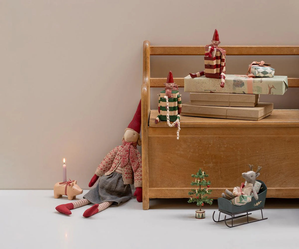 A Maileg Elf with Drawer in a red outfit (pre-order, arriving mid August), sits on a wooden bench with gift boxes, beside a festive candle in a pig holder, toy reindeer in sleigh, and mini Christmas tree on a white floor.