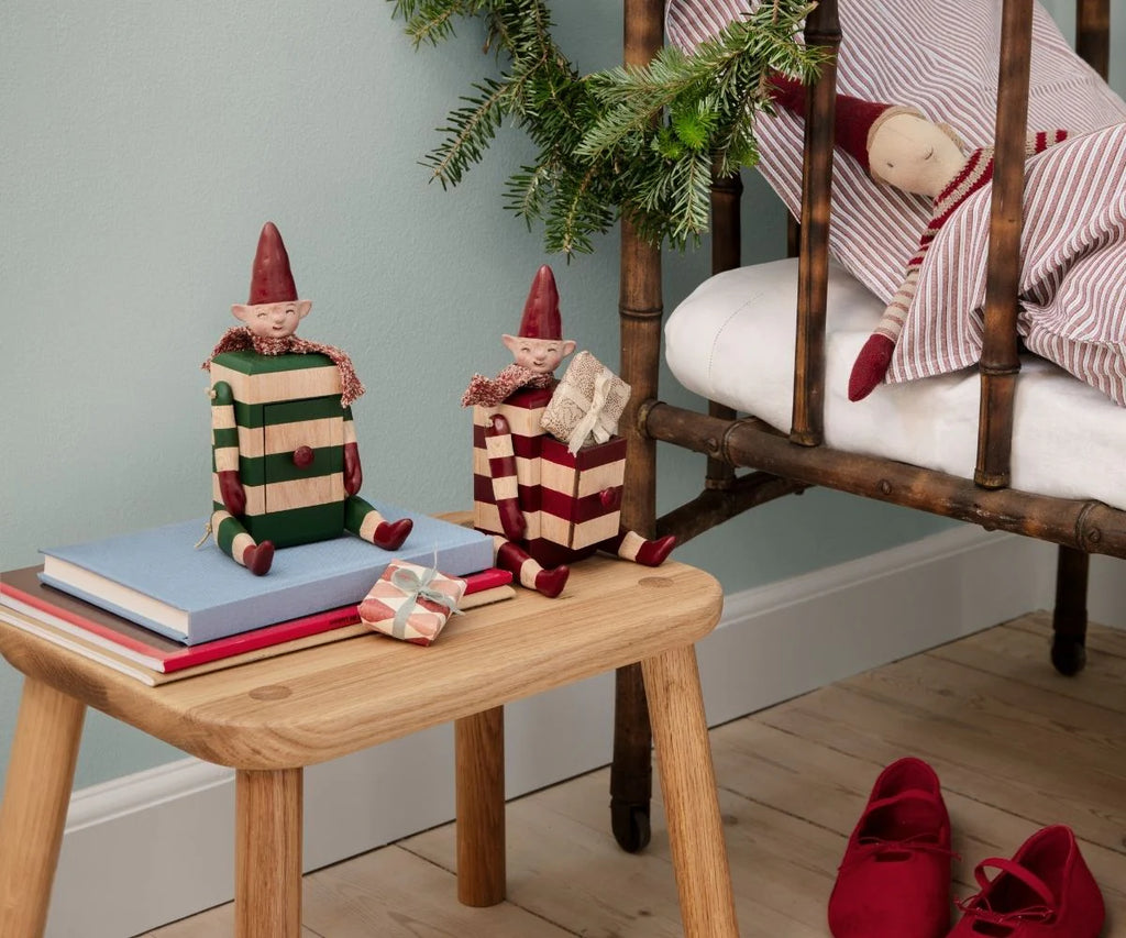 A Maileg Elf with Drawer - Green (pre-order, arriving mid August) sits on a wooden stool near a gift box and books, while another elf in red and white rests on a rustic bed with red shoes and a pine branch nearby.