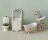 The Maileg Beach Mouse With Float, Little Sister - Red Dot, wearing glasses and a tiny swimsuit with a red dotted swim ring, sits on a stool beside a toy bathtub in a miniature bathroom playset complete with toy toilet and vanity, all set against charming patterned wallpaper for an adorable scene.