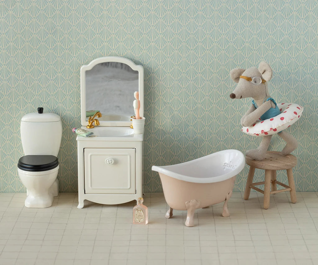 The Maileg Beach Mouse With Float, Little Sister - Red Dot, wearing glasses and a tiny swimsuit with a red dotted swim ring, sits on a stool beside a toy bathtub in a miniature bathroom playset complete with toy toilet and vanity, all set against charming patterned wallpaper for an adorable scene.