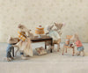 Six Maileg mice from the adorable mice family gather around a table with the Maileg Party Cake and Cake Stand, Mouse - Off White, as one mouse stands on a chair pouring syrup. A wrapped gift rests beneath the table, while all the mice are dressed in cute, colorful outfits.