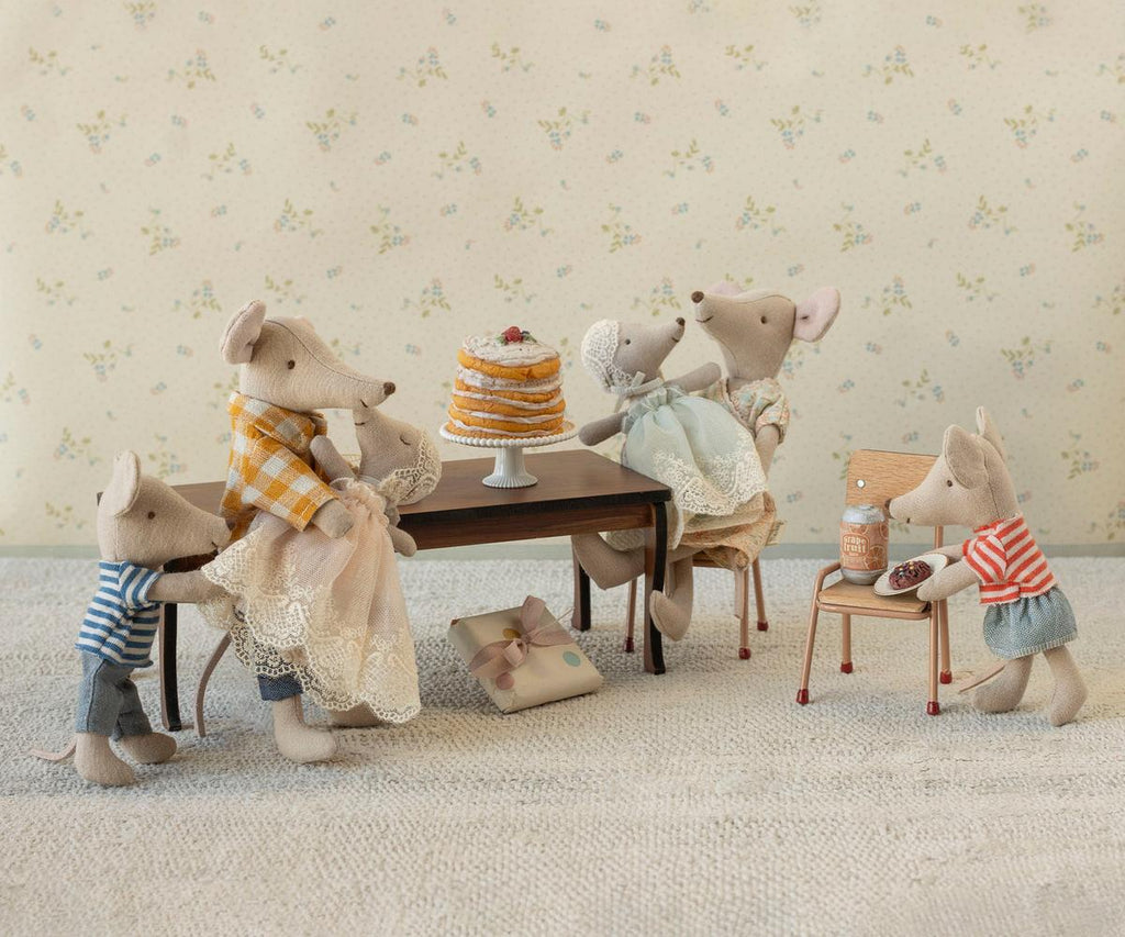 Five plush mice gather around the Maileg Long Dining Table, Mouse, which is topped with a tall layered cake. One mouse stands on a chair, preparing tea, while a wrapped gift sits on the floor. The scene feels cozy and whimsical, enhanced by charming dollhouse accessories and soft pastel tones.