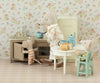Three stuffed toy mice are in a vintage-style dollhouse kitchen. One mouse opens an oven, another stands at a Maileg Vintage Dining Table, Mouse - Off White, with bread and dishes, and a third sits in a highchair. Floral wallpaper and dollhouse furniture, including a toy fridge, are in the background.