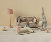 Three Maileg Rabbit With Accessories, Micro - Sand - Rose toys are in a miniature living room with a beige couch, floral rug, rocking chair, lamp, ball, and teacup. One bunny rolls out the rug while the others look on in this charming scene.