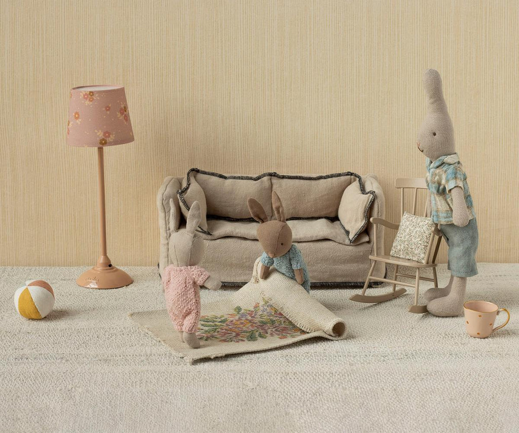 Three Maileg Rabbit With Accessories, Micro - Sand - Rose toys are in a miniature living room with a beige couch, floral rug, rocking chair, lamp, ball, and teacup. One bunny rolls out the rug while the others look on in this charming scene.