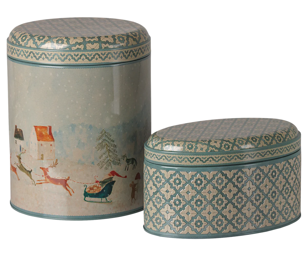 PRE-ORDER: Maileg Metal Box Set - Santa is Coming includes two decorative Christmas boxes—one cylindrical, one oval. Both feature blue and gold patterned lids and borders; the larger metal box shows Santa’s sleigh with reindeer in a snowy scene.