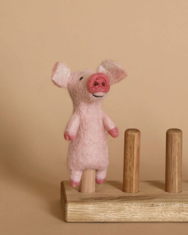 The Pig Finger Puppet, a delightful small pink needle-felted creation with a smiling face, perches on one of three wooden pegs, serving as an enchanting handmade storytelling prop against the neutral beige background.