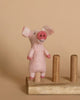 The Pig Finger Puppet, a delightful small pink needle-felted creation with a smiling face, perches on one of three wooden pegs, serving as an enchanting handmade storytelling prop against the neutral beige background.