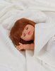 A small Minikane Baby Doll With Sleeping Eyes (13") - Capucine with straight brown hair and closed eyes is wrapped snugly in a white knitted blanket. The doll has a peaceful expression, its right hand slightly raised, and exudes a natural vanilla scent against a soft, knitted white fabric background.