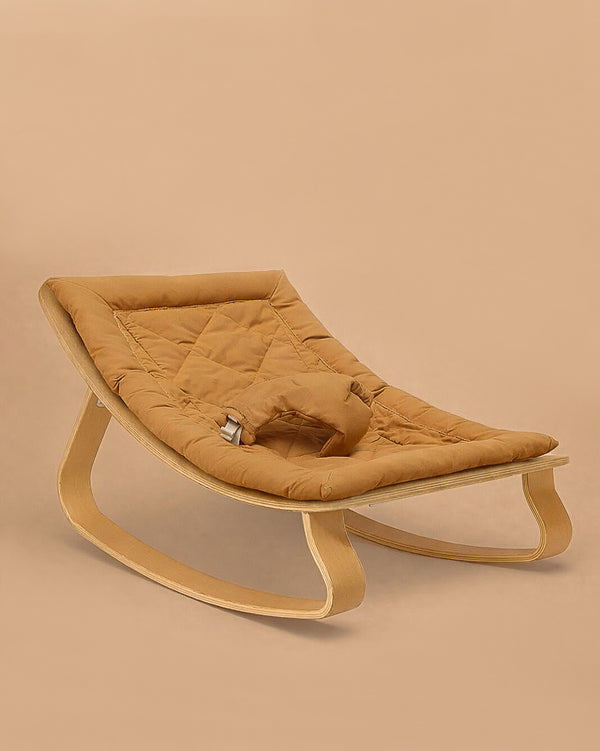 The Charlie Crane LEVO Baby Rocker features a light wooden frame and a padded camel cushion, shown here against a matching tan background.