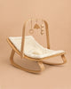 The Charlie Crane LEVO Baby Rocker with Organic White Cushion features a soft, white seat and a minimalistic toy bar with wooden hanging toys, shown against a beige background.