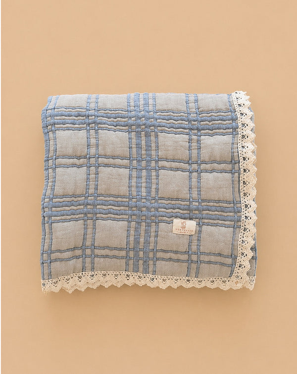 The Organic Cotton Baby Quilt - French Plaid, Bluebelle is a 30x40 inch blue checkered quilt with white lace trim, made from organic cotton in Portugal and shown on a beige background.