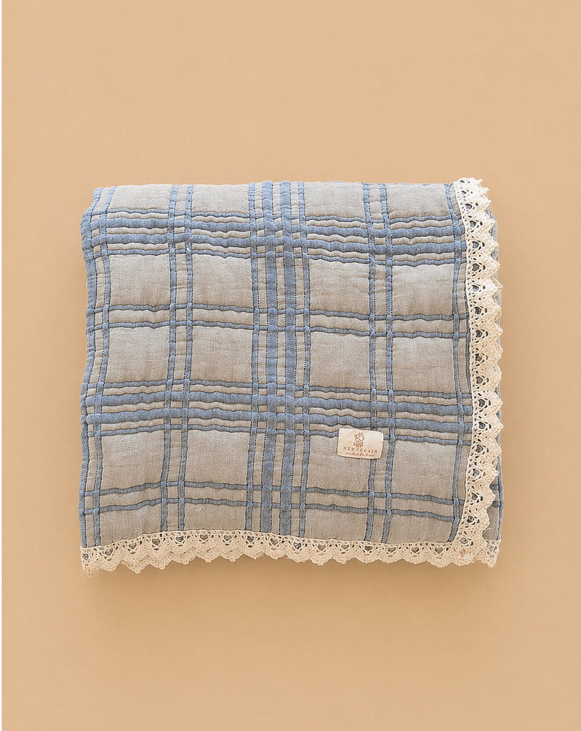 The Organic Cotton Baby Quilt - French Plaid, Bluebelle is a 30x40 inch blue checkered quilt with white lace trim, made from organic cotton in Portugal and shown on a beige background.