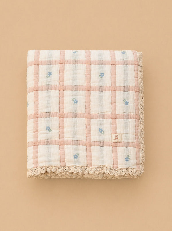 The Organic Cotton Baby Quilt - Meadow, Petal, made in Portugal, features a pink and white checkered design with delicate blue flowers and lace trim, displayed on a neutral beige background.