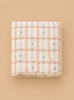 The Organic Cotton Baby Quilt - Meadow, Petal, made in Portugal, features a pink and white checkered design with delicate blue flowers and lace trim, displayed on a neutral beige background.